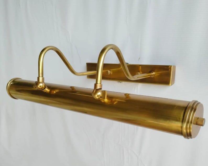 May include: A brass picture light fixture with a rectangular wall mount and a cylindrical light housing. Two curved arms extend from the housing. The fixture is a warm gold color.