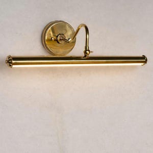 May include: A brass picture light fixture with a circular wall mount and a curved arm. The light bar is horizontal and has a bright, warm glow. The fixture is mounted on a light-colored wall.