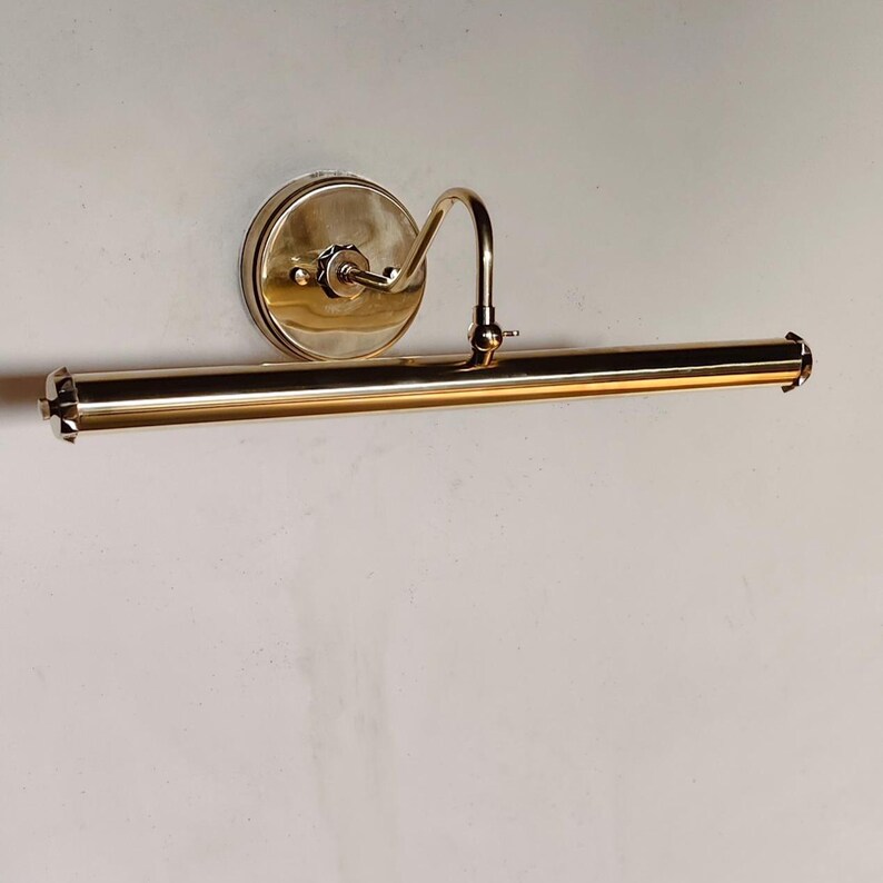 May include: A brass-toned picture light fixture. The fixture has a circular wall mount, a curved arm, and a long, cylindrical light bar. The light bar has decorative end caps. The fixture is mounted on a white wall.