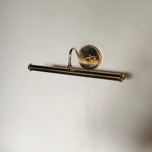 May include: A brass picture light fixture with a curved arm and a circular backplate. The light has a rectangular bar with decorative end caps. The fixture is mounted on a white wall.