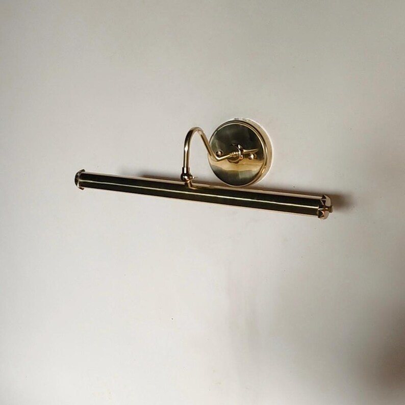 May include: A brass picture light fixture with a curved arm and a circular backplate. The light has a rectangular bar with decorative end caps. The fixture is mounted on a white wall.