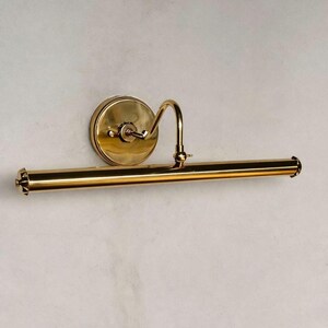 May include: A brass picture light fixture with a curved arm and a round backplate. The light has a cylindrical bar and decorative end caps. The fixture is mounted on a white wall.