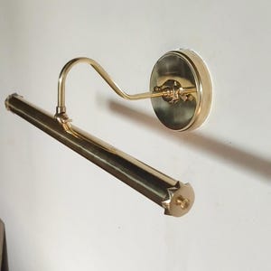 May include: A brass picture light fixture with a curved arm and a cylindrical light bar. The fixture is mounted on a round backplate. The light bar has a starburst design on the end.