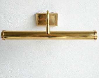 May include: A brass picture light fixture with a rectangular backplate and a long, cylindrical light bar. The fixture is mounted on a textured white wall. The light bar is approximately 24 inches long.