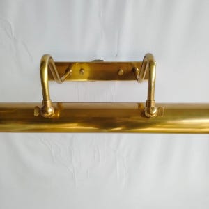 May include: A vintage brass picture light fixture. The fixture has a long, cylindrical body with a curved handle on each end. The mounting plate has two curved arms. The brass has a polished finish.