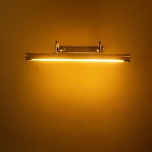 May include: A brass-toned picture light fixture with a bright, warm yellow glow. The light is mounted on a wall and has a rectangular shape with a curved tube. The fixture is designed to illuminate artwork.