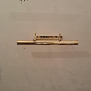 May include: A brass picture light fixture. The fixture has a long, cylindrical light housing with a curved bracket and a rectangular mounting plate. The light is mounted on a neutral-colored wall.