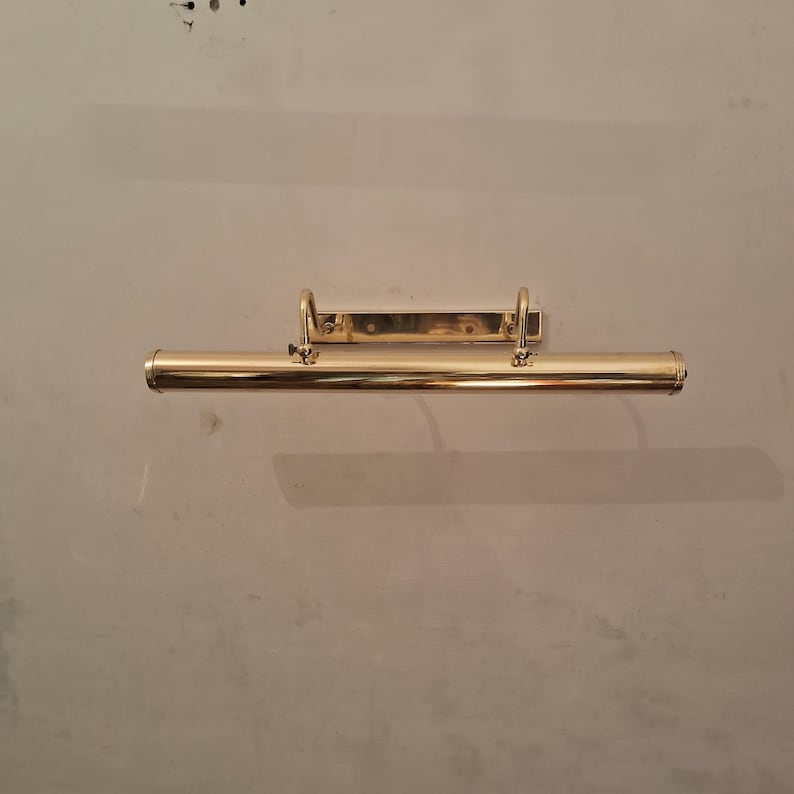 May include: A brass picture light fixture. The fixture has a long, cylindrical light housing with a curved bracket and a rectangular mounting plate. The light is mounted on a neutral-colored wall.