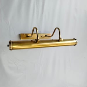 May include: A brass picture light fixture with a rectangular backplate and a cylindrical light housing. Two curved arms extend from the backplate to support the light housing. The fixture is a warm gold color.