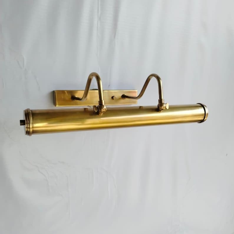 May include: A brass picture light fixture with a rectangular backplate and a cylindrical light housing. Two curved arms extend from the backplate to support the light housing. The fixture is a warm gold color.