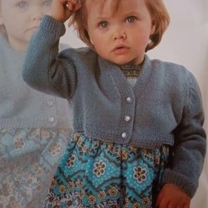 May include: A young child wearing a blue knitted cardigan with three white buttons. The child is also wearing a blue and floral patterned dress. The image has a soft, vintage aesthetic, with a blurred reflection of the child in the background.
