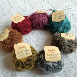 Può includere: Una collezione di gomitoli di lana Katia Danubio Style in vari colori, tra cui rosso, viola, verde acqua, marrone, giallo e grigio. Ogni gomitolo ha un'etichetta con il nome del marchio e lo stile. La lana è disposta in un motivo circolare su una superficie bianca strutturata.