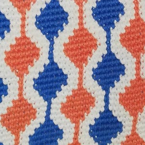 Puede incluir: Primer plano de una tela tejida con un patrón repetitivo. El diseño presenta formas de diamante en azul y naranja, separadas por líneas blancas. La textura de la tela es visible, con cada puntada claramente definida.