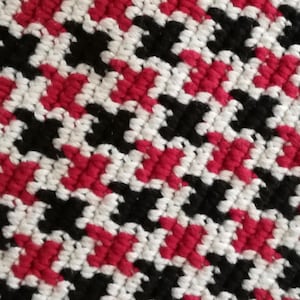 May include: Close-up of a houndstooth textile. The pattern is a repeating design of black, white, and red shapes. The fabric has a woven or knitted appearance, with a textured surface.