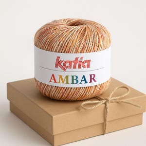 Puede incluir: Ovillo de lana Katia Ambar en tonos naranja, beige y crema, sobre una caja de cartón marrón atada con hilo. La etiqueta de la lana presenta el logotipo de Katia y la palabra "AMBAR" en colores arcoíris.