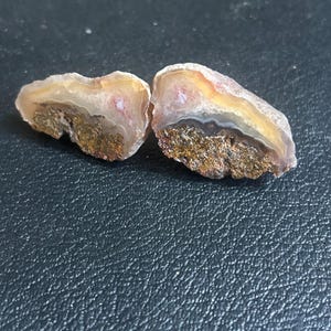 May include: Two polished agate stones, each displaying a unique pattern of colors. The stones feature bands of beige, brown, and white, with hints of pink and yellow. The bottom portion of each stone has a rough, textured appearance.