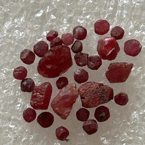 May include: A collection of raw, uncut ruby gemstones in varying sizes and shapes. The rubies display a deep red hue, with some stones appearing more translucent than others. The gemstones are scattered on a textured, white surface.