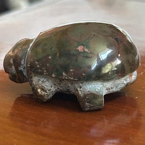 May include: A small, bronze-colored turtle figurine with a glossy shell and textured underside. The turtle's shell has a mottled appearance, with hints of green and brown. The figurine is resting on a wooden surface.