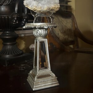 May include: A silver-colored decorative pedestal with a rectangular mirrored base and a round top supporting a large, white crystal geode. The geode is a cluster of small, sparkling crystals.