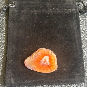 May include: An orange and white agate stone rests on a black velvet pouch. The agate has a polished surface with concentric bands of color. The pouch has a drawstring closure.
