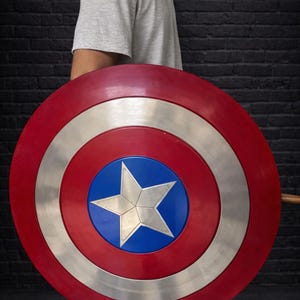 May include: A red, white, and blue shield with a star in the center. The shield has a circular design with a silver ring and a blue center. The shield is held by a person wearing a gray t-shirt.