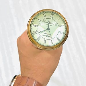 May include: A brass pocket watch with a cream face and black Roman numerals. The watch face features the text "Regal Clocks Co. Est 1840" and "Scotland". The watch is held in a hand.