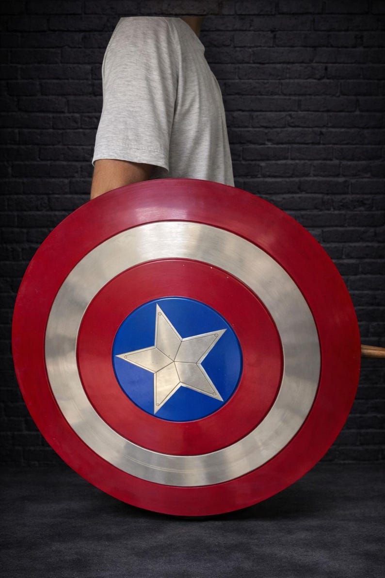 May include: A red, white, and blue shield with a star in the center. The shield has a circular design with a silver ring and a blue center. The shield is held by a person wearing a gray t-shirt.
