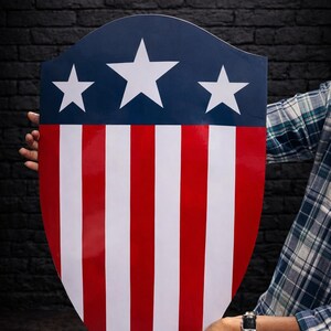 May include: A shield with a patriotic design. The top section is navy blue with three white stars. The shield features alternating red and white vertical stripes, creating a bold and iconic look. The shield is held by a person.