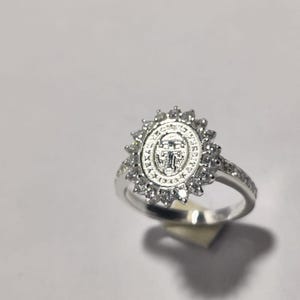 May include: A silver ring with a central oval design surrounded by a sunburst of small, clear stones. The ring band is also adorned with small stones. The ring is set against a white background.