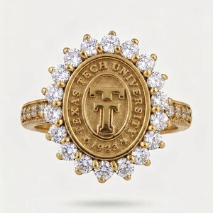 May include: A gold ring featuring an oval centerpiece with the embossed text Texas Tech University and 1923 surrounding a central TT logo. The oval is framed by a halo of clear round stones, with additional stones set along the gold band.