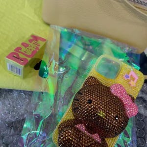 May include: A yellow phone case with a bedazzled Hello Kitty design, featuring a pink bow and star accents. A small box of "DREAM LIP BALM" is visible, along with a yellow bag and iridescent packaging.