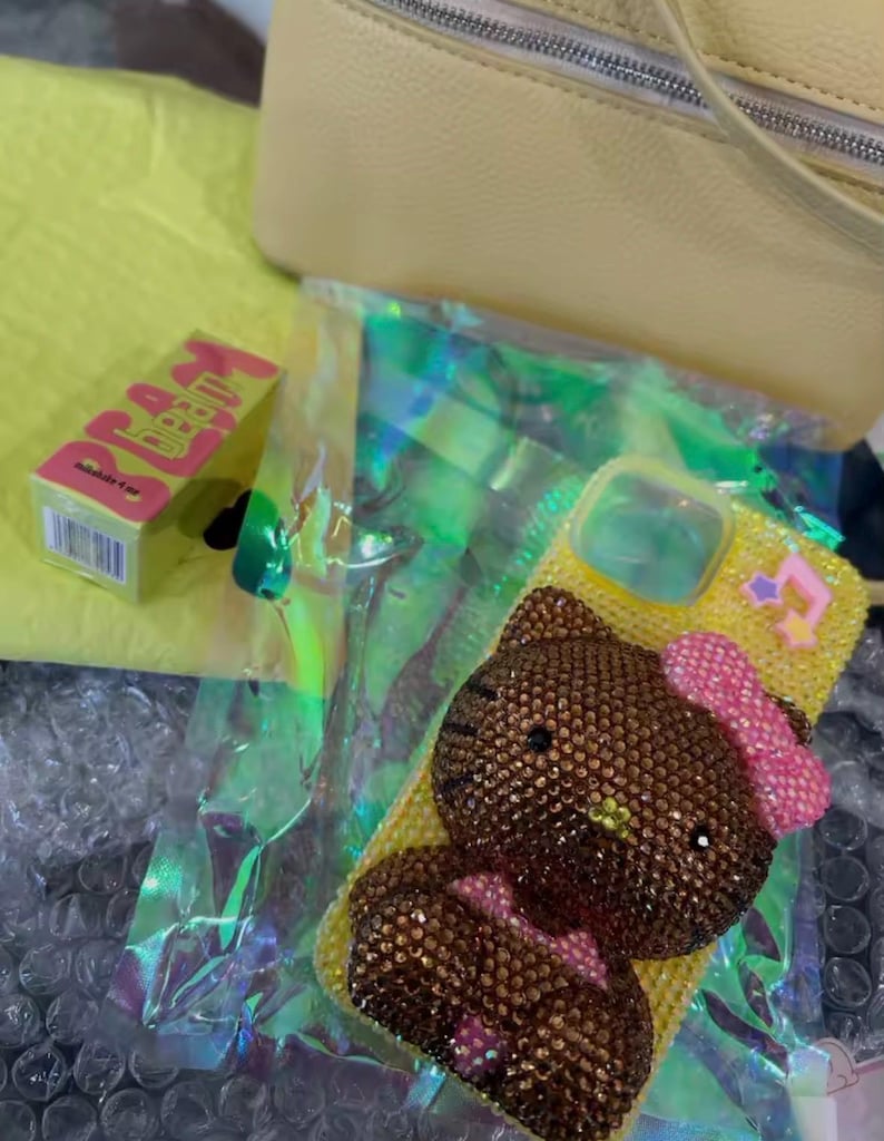 May include: A yellow phone case with a bedazzled Hello Kitty design, featuring a pink bow and star accents. A small box of "DREAM LIP BALM" is visible, along with a yellow bag and iridescent packaging.