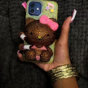 May include: A bedazzled phone case featuring a brown Hello Kitty design with a pink bow. The case is covered in small, sparkling rhinestones. The phone case is held in a hand with long, manicured nails and gold bracelets.