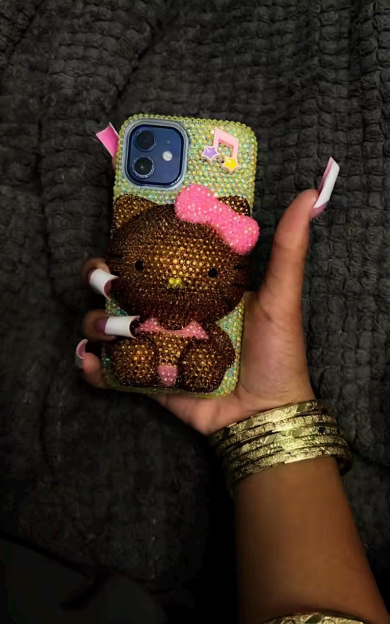 May include: A bedazzled phone case featuring a brown Hello Kitty design with a pink bow. The case is covered in small, sparkling rhinestones. The phone case is held in a hand with long, manicured nails and gold bracelets.