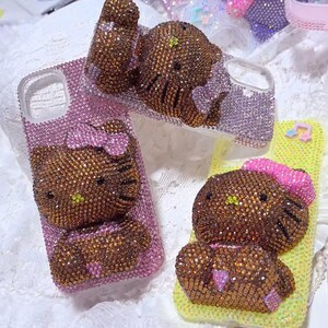 May include: Several bedazzled phone cases featuring a brown Hello Kitty design. The cases are covered in sparkling rhinestones in shades of pink, purple, and yellow. Each case has a different Hello Kitty pose, with bows and other details.