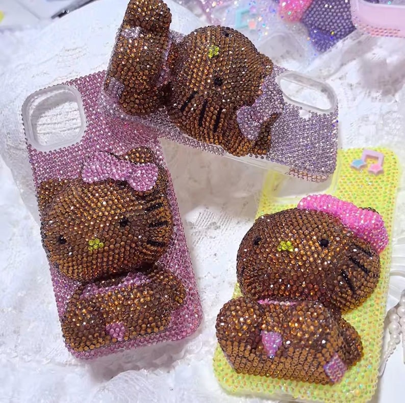 May include: Several bedazzled phone cases featuring a brown Hello Kitty design. The cases are covered in sparkling rhinestones in shades of pink, purple, and yellow. Each case has a different Hello Kitty pose, with bows and other details.