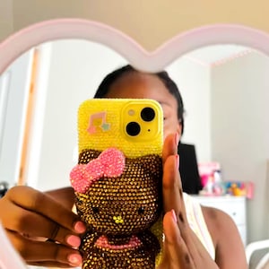 May include: Yellow phone case with a Hello Kitty design, embellished with rhinestones. The case features a pink bow and musical note accents. The phone case is held up in front of a mirror.