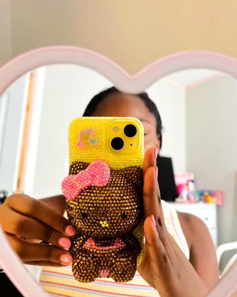 May include: Yellow phone case with a Hello Kitty design, embellished with rhinestones. The case features a pink bow and musical note accents. The phone case is held up in front of a mirror.