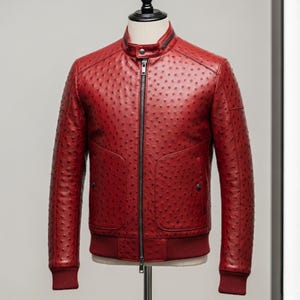 Può includere: Giacca in pelle rossa con superficie strutturata e chiusura a zip. La giacca ha un collo alto, due tasche anteriori e polsini e orlo a coste. La giacca è esposta su un manichino.