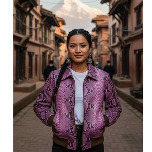 Peut inclure: Une femme porte une veste rose à motif peau de serpent avec un t-shirt blanc en dessous. La veste a une fermeture éclair et des bordures marron. Elle se tient dans une rue pavée avec des bâtiments de chaque côté.