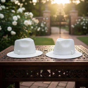 May include: Two white fedora hats with a textured pattern and a band around the crown sit on a dark wooden table. The hats are displayed outdoors with a garden and brick wall in the background.