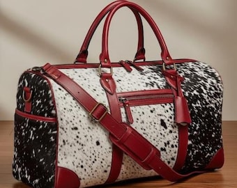 Könnte beinhalten: Reisetasche aus Kuhfell mit schwarz-weißem Fleckenmuster. Die Tasche hat rote Lederakzente an den Griffen, der Einfassung und dem Schulterriemen. Sie verfügt über eine Reißverschluss-Vordertasche und einen passenden Lederanhänger.