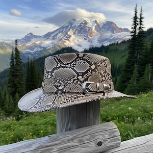 Może przedstawiać: Kapelusz fedora z wzorem skóry węża ze srebrną klamrą i paskiem. Kapelusz jest prezentowany na drewnianym słupku z pasmem górskim w tle. Wzór kapelusza to mieszanka szarości, brązu i bieli, z szerokim rondem.