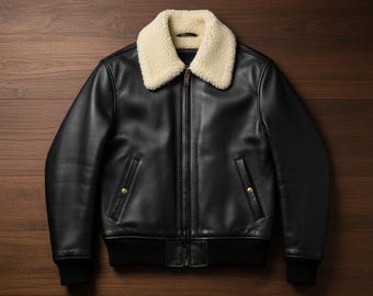 May include: Black leather jacket with a cream-colored shearling collar. The jacket features a zip-up front, two front pockets with gold-colored button closures, and black ribbed cuffs and hem. The jacket is displayed on a wooden surface.