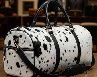 Könnte beinhalten: Zylindrische Reisetasche mit weißem und schwarzem Fleckenmuster. Sie hat schwarze Ledergriffe, Riemen und einen Schultergurt. Die Tasche verfügt über einen Reißverschluss und goldfarbene Metallbeschläge.