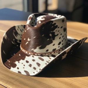 May include: Cowboy hat with a brown and white spotted pattern. The hat features a brown leather band around the crown and a wide brim. The hat is made of a soft, textured material, and is likely made of animal hide.