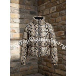 Puede incluir: Chaqueta acolchada con estampado de piel de serpiente en tonos marrón, beige y negro. La chaqueta tiene cuello alto, cremallera completa y corte corto. Sobre un fondo de pared de ladrillo.