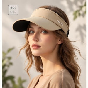 May include: A beige sun visor featuring a wide brim with a black underside and the text UALACL printed on the side. The visor includes a square graphic in the corner displaying the text UPF 50+ to indicate sun protection capabilities.