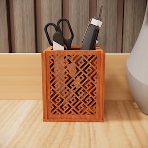 Op de afbeelding: Een bruine houten bureau-organizer met een geometrisch patroon, met een schaar en een pen. De organizer staat op een lichtgekleurd houten oppervlak. Een witte en grijze vaas staat op de achtergrond.