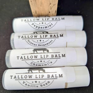 May include: Four white lip balm tubes with labels. Each label reads "TALLOW LIP BALM" above a flavor name: Peppermint, Orange Bergamot, Chamomile, and Vanilla Bean. A small illustration of a cow is on each label.